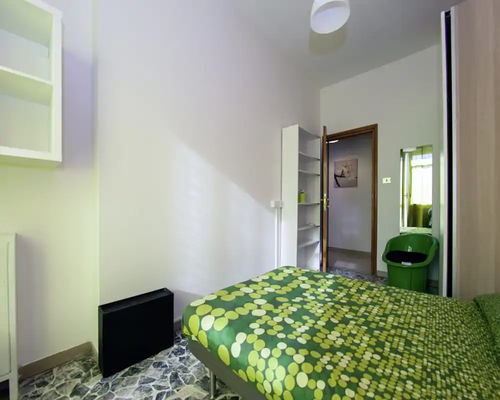 Chambre compacte avec lit double vert à motifs, étagères intégrées et lumière naturelle. La pièce est propre, bien éclairée et adaptée à la location courte durée.