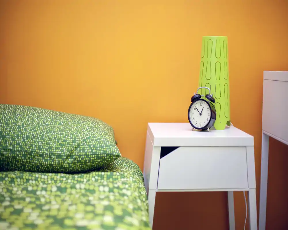 Coin de chambre lumineux et coloré avec un lit vert à motif et une table de chevet blanche surmontée d'un réveil rétro et d'une lampe décorative; le mur orange chaud crée une ambiance cosy et moderne.