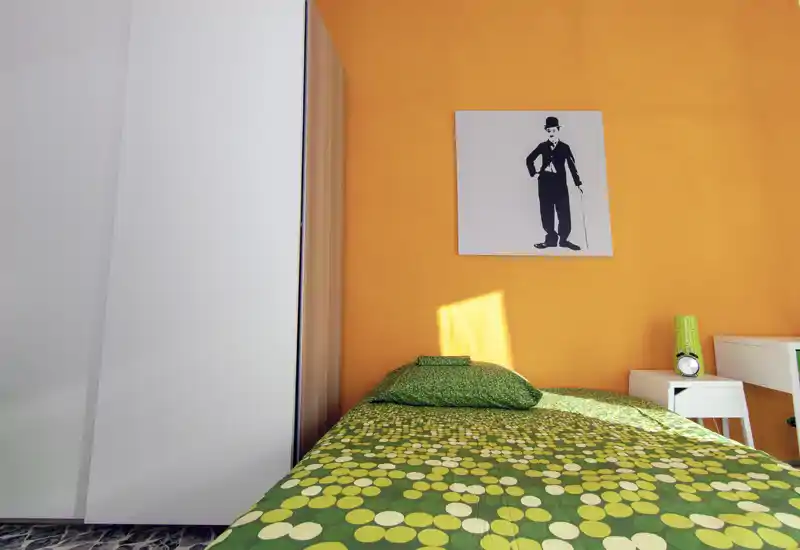 Camera da letto singola luminosa con copriletto verde a motivo e parete arancione; comodino, lampada e armadio sono visibili. La stanza è fresca e ben illuminata, ideale per un annuncio accogliente.
