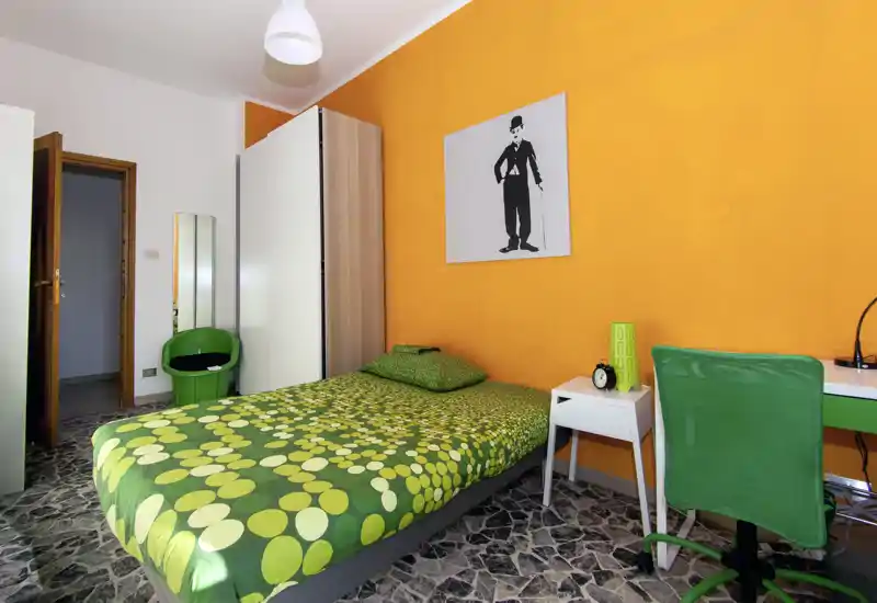 Camera singola luminosa e colorata con letto verde a fantasia, comodino e scrivania. La parete arancione e i tessuti vivaci creano un’atmosfera accogliente ideale per studenti o affitti brevi.