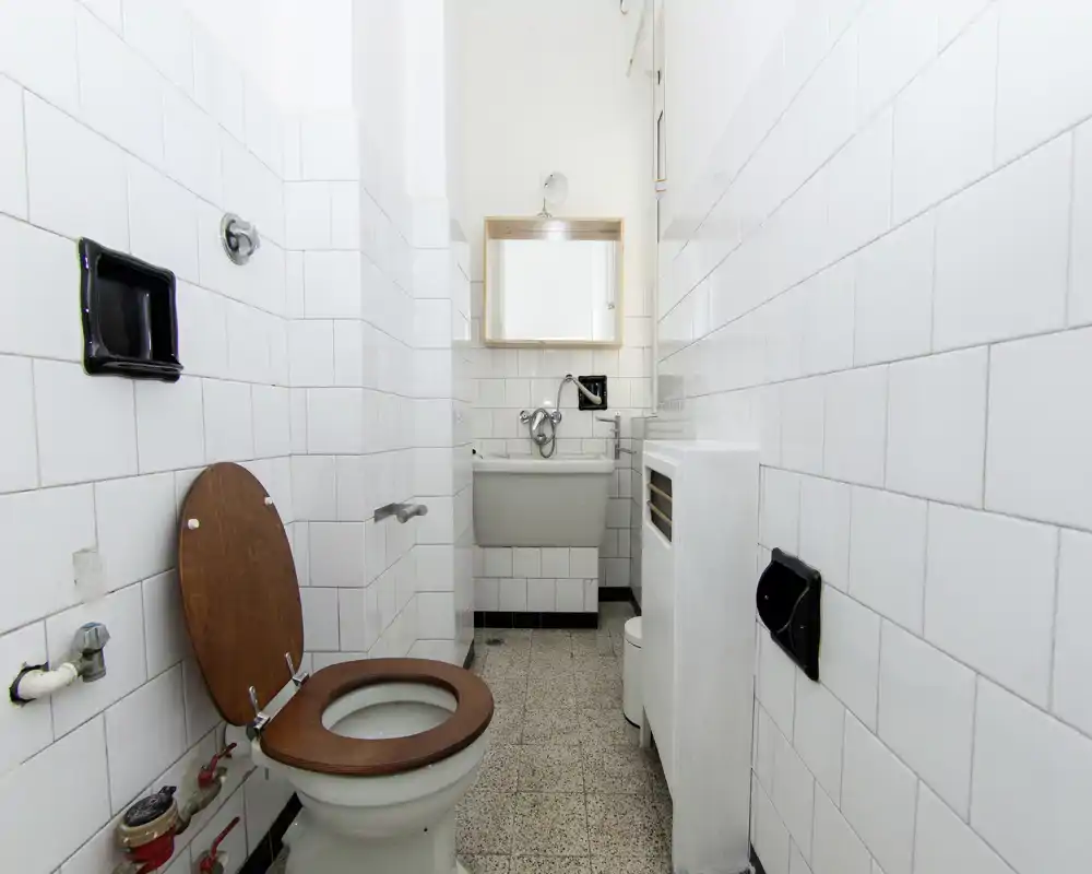 Petite salle de bain carrelée avec WC à siège en bois, lavabo mural et miroir ; lumineuse, fonctionnelle et bien photographiée.