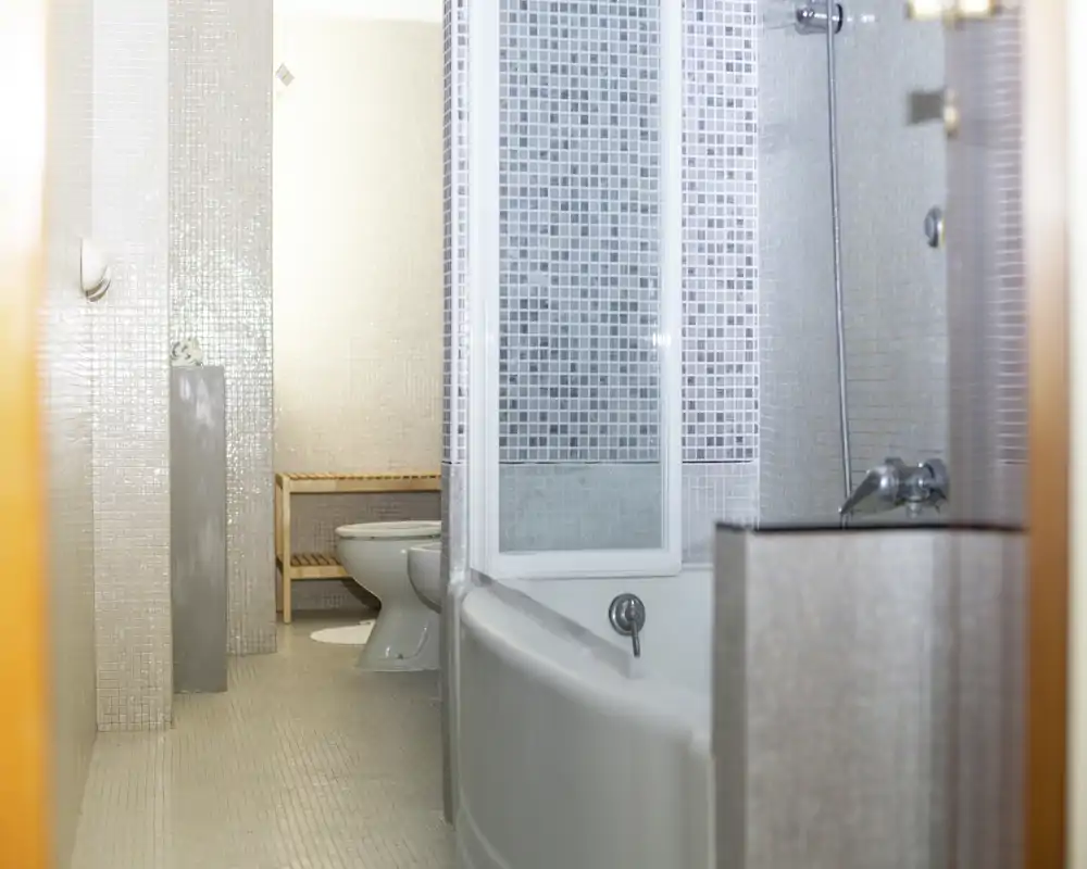 Salle de bains lumineuse carrelée avec baignoire et pare-douche, WC et petit banc; carreaux mosaïque neutres et bonne lumière naturelle.