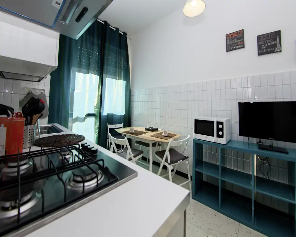 Cuisine compacte et lumineuse avec plaque à gaz au premier plan, petite table pour quatre et étagère bleu-vert. La lumière de la fenêtre et les textiles harmonieux offrent un espace pratique et chaleureux, parfait pour des séjours courts.