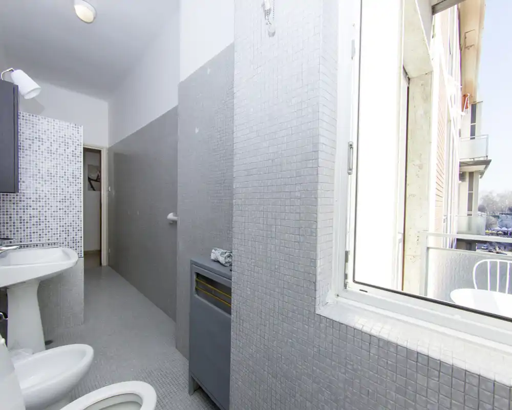 Salle de bain carrelée et lumineuse avec bidet, WC et lavabo sur colonne ; une grande fenêtre donne sur un petit balcon et apporte beaucoup de lumière naturelle.