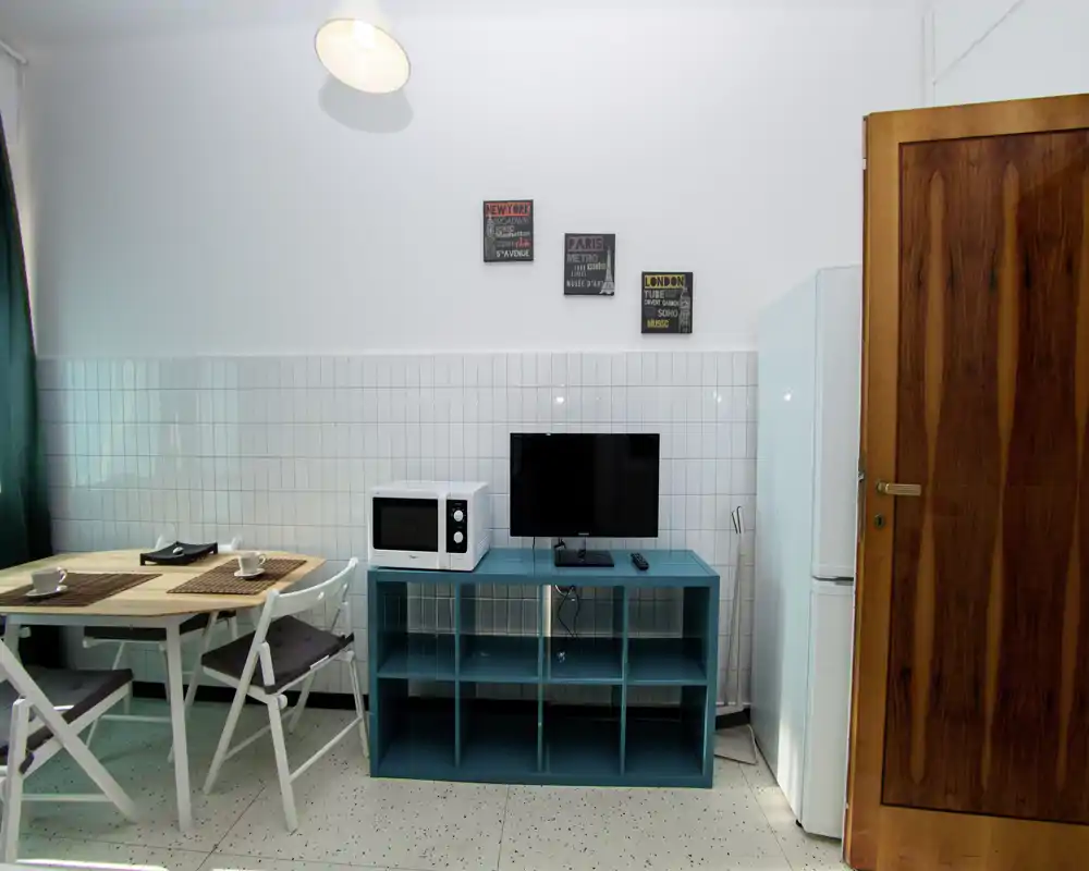 Coin cuisine compact avec meuble de rangement turquoise, micro-ondes, petite télévision, réfrigérateur et une table à manger simple avec deux chaises. Espace lumineux et bien rangé, décoré de plaques murales et de rideaux verts.