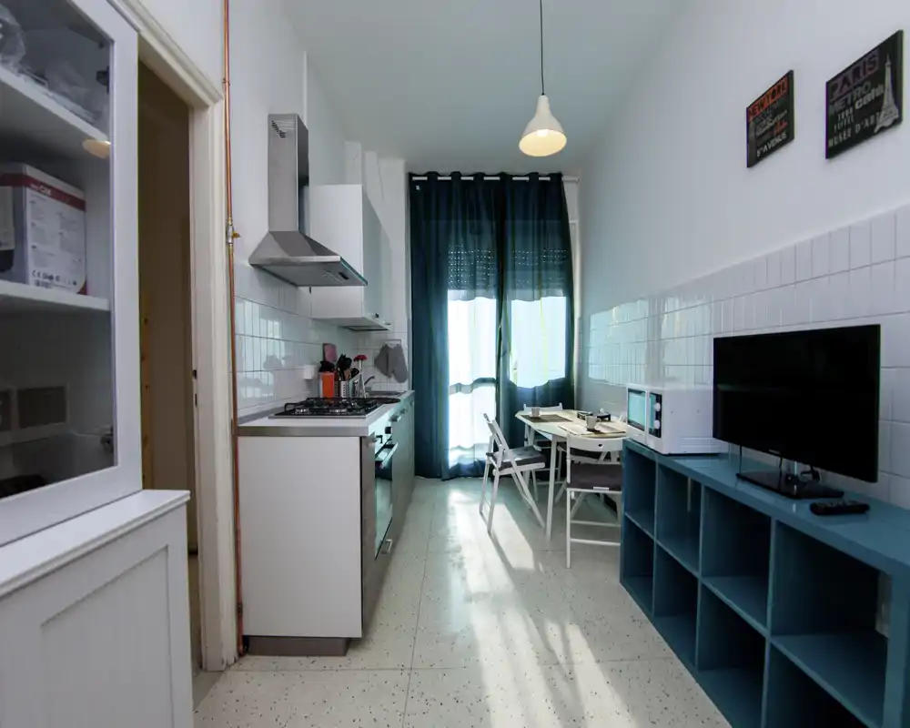 Kitchenette moderne et lumineuse avec plaque de cuisson intégrée, hotte et petit coin repas près d'une porte-fenêtre ; accents turquoise et étagères coordonnées offrent une ambiance fraîche et accueillante.