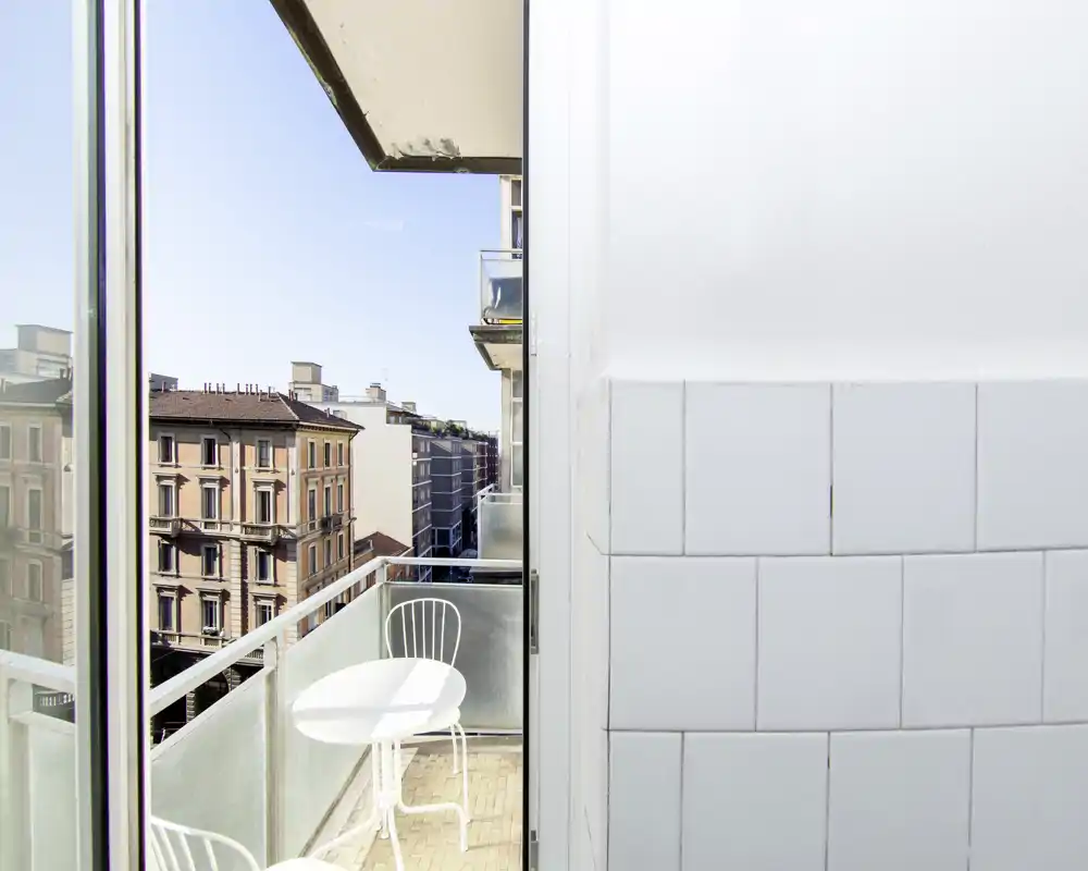Petit balcon ensoleillé avec table et chaise blanches, vue sur des immeubles urbains classiques — espace extérieur lumineux idéal pour un café matinal.