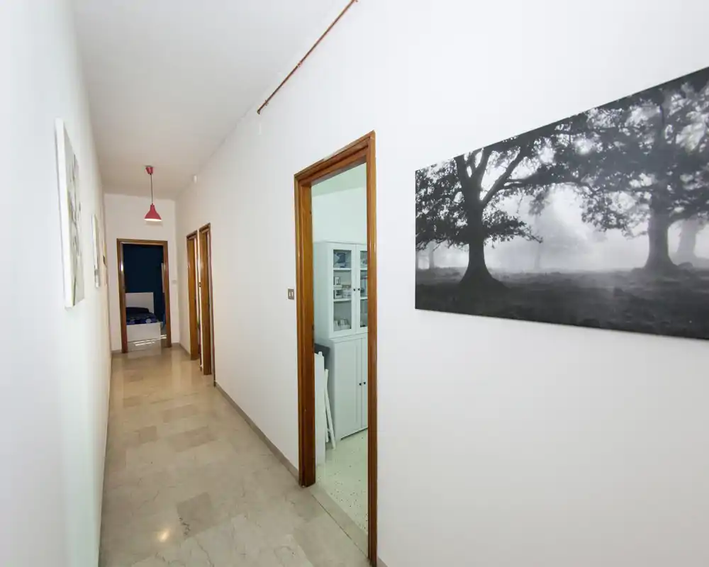 Couloir intérieur lumineux et long avec sol carrelé, murs blancs et encadrements de porte en bois menant à plusieurs pièces ; décoration simple et tableau mural.