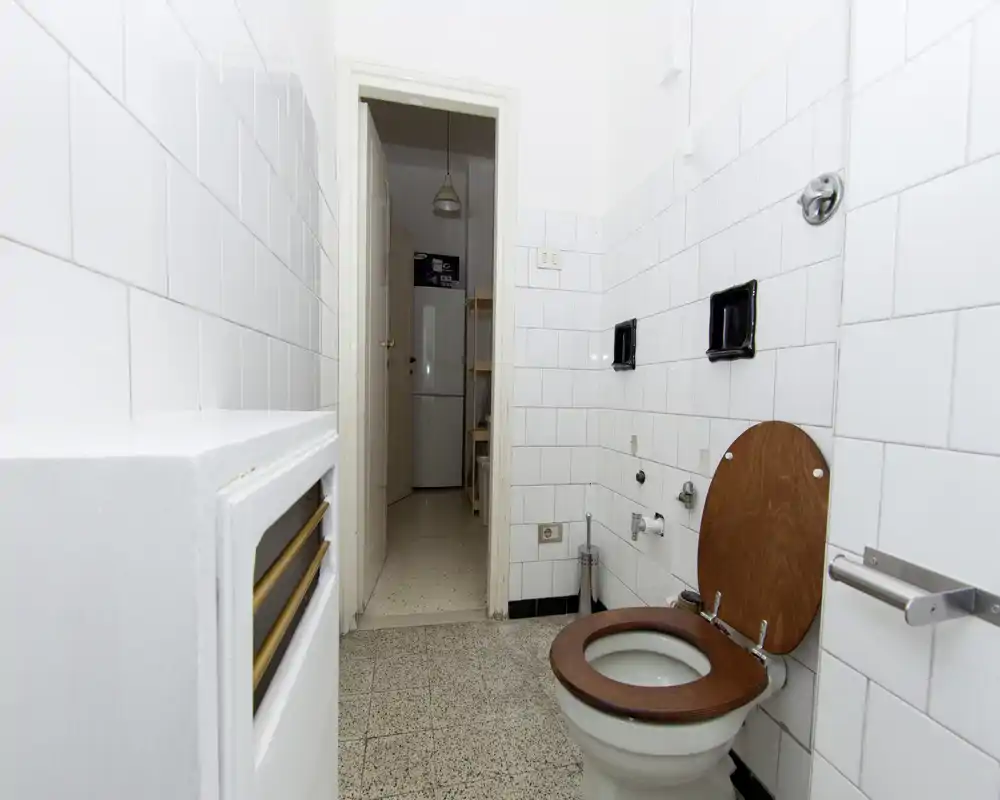 Petite salle de bains carrelée de blanc avec WC à lunette en bois, murs carrelés et vue par la porte sur un couloir menant à la cuisine.