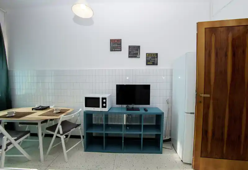 Angolo cucina compatto con mobiletto turchese, microonde, piccolo televisore, frigorifero e un semplice tavolo da pranzo con due sedie. Spazio luminoso e ordinato con targhette decorative e tende verdi.