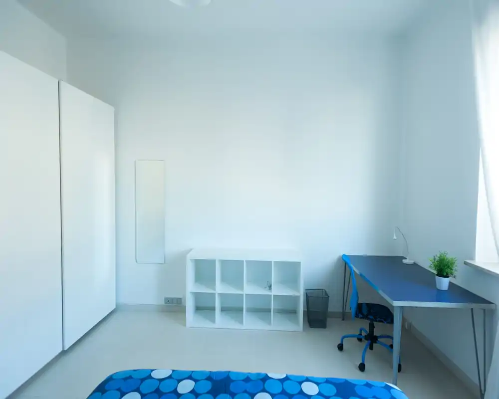 Camera da letto luminosa e minimalista con vista parziale del letto, grande armadio, scaffalatura e una scrivania vicino alla finestra — ordinata e ariosa.