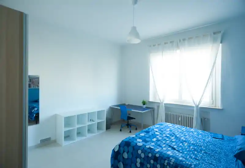 Camera da letto luminosa e ordinata con copriletto blu a motivi, scrivania vicino a una grande finestra e scaffalatura minimal—ideale per uno studente o una persona sola.