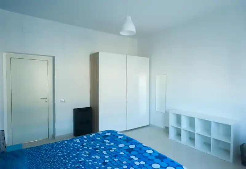 Camera da letto luminosa e minimalista con copriletto blu a pois, ampia armadiatura bianca e scaffalatura aperta; linee pulite e buona luce naturale, ideale per annunci.