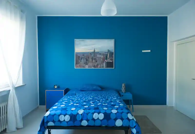 Camera da letto luminosa e moderna con parete di accentuazione blu e biancheria a fantasia blu. La luce naturale da una grande finestra e i semplici comodini creano un ambiente fresco e confortevole.