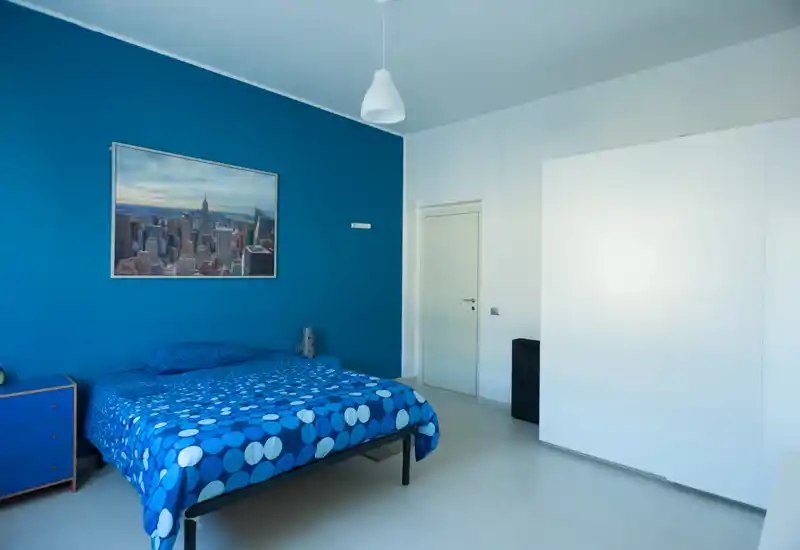 Camera da letto moderna e luminosa con letto matrimoniale contro una parete blu, comodino semplice e un grande armadio bianco. Linee pulite, arredo contemporaneo e luce naturale rendono l’ambiente fresco e arioso.