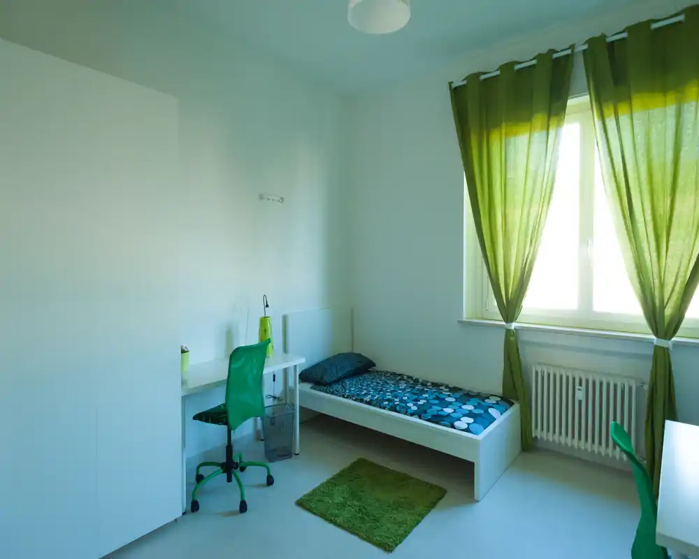 Camera da letto luminosa e arredata in modo essenziale con letto singolo, scrivania e armadio. Dettagli verdi (tende, sedia e tappeto) conferiscono un aspetto fresco e coordinato, con abbondante luce naturale.