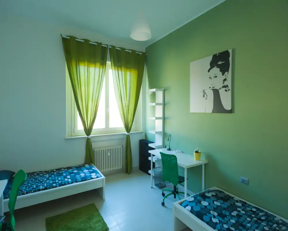 Camera doppia luminosa con due letti singoli, dettagli verdi e una scrivania vicino alla finestra. Luce naturale, tessuti coordinati e arredo minimal moderno creano uno spazio fresco e confortevole, ideale per studenti o soggiorni brevi.