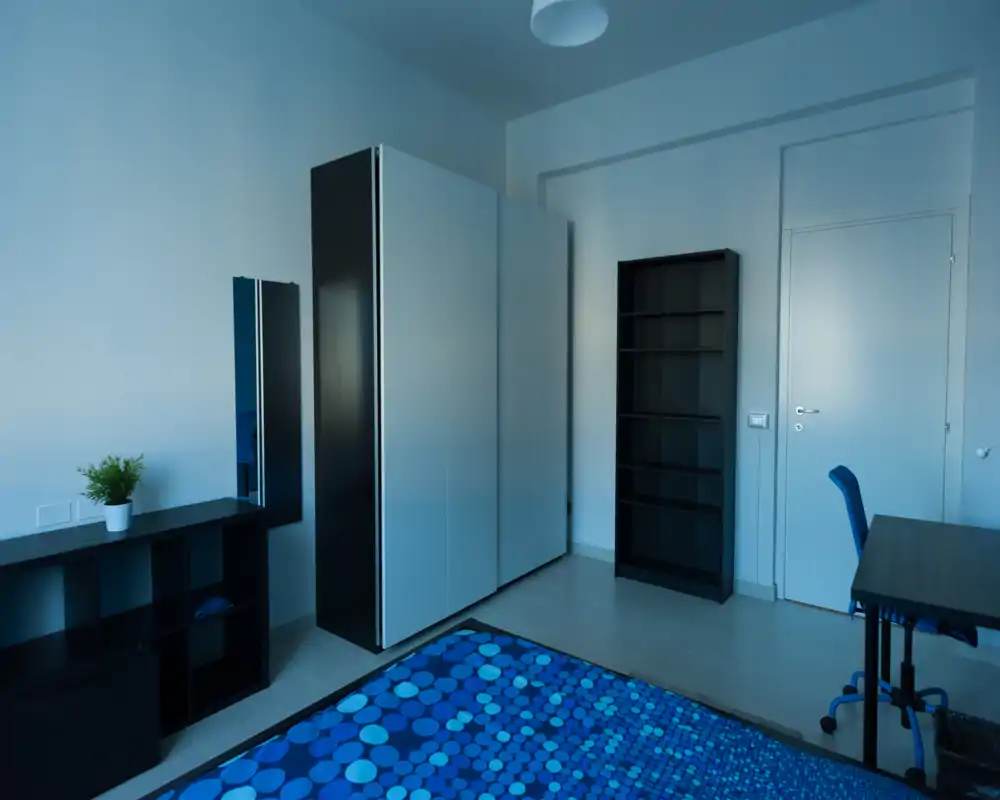 Camera da letto moderna con copriletto blu a motivi, armadio alto bianco e scaffalature. Arredamento pulito e minimale con scrivania e sedia, ideale per una singola occupazione comoda.