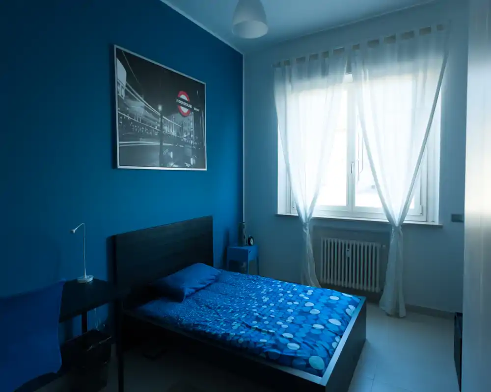 Camera da letto dallo stile moderno in tonalità blu, con letto matrimoniale, comodino e ampia finestra che filtra luce naturale soffusa.