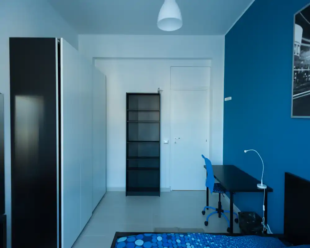 Camera da letto moderna e luminosa con parete blu, letto matrimoniale con biancheria blu a motivo, armadio, scrivania e scaffale — ideale per studenti o professionisti che cercano uno spazio ordinato e funzionale.