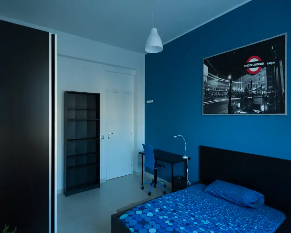 Camera da letto moderna con copriletto blu a motivo, testiera semplice e scrivania vicino alla parete. Parete accentuata in blu, libreria e lampada a sospensione dal design contemporaneo.