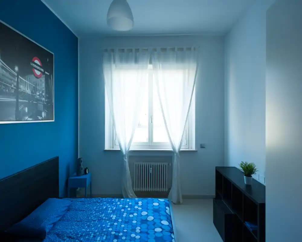 Camera da letto luminosa e moderna con parete blu e biancheria a fantasia, tende leggere che filtrano la luce e un semplice modulo contenitore per oggetti.