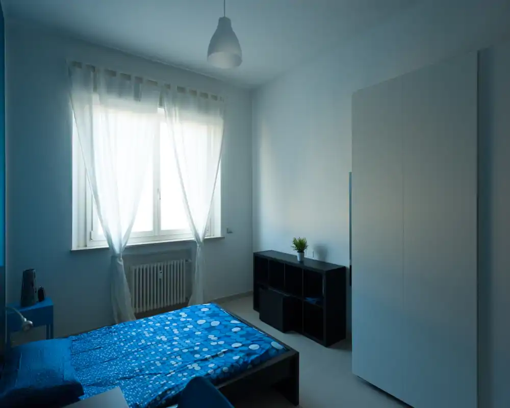 Camera da letto luminosa e arredata in modo semplice con letto matrimoniale, copriletto blu a fantasia, armadio e mobile con scomparti; la luce naturale dalle tende leggere crea un’atmosfera tranquilla.