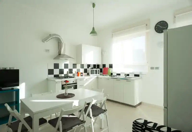 Cucina moderna e luminosa con tavolo da pranzo: mobili bianchi, schienale a scacchi, elettrodomestici e una finestra soleggiata che rendono l'ambiente arioso e accogliente.