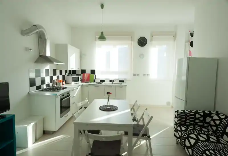 Angolo cottura moderno e luminoso con un piccolo tavolo da pranzo e quattro sedie, piano cottura a gas, forno, microonde e frigorifero. Lo spazio è pulito, ben illuminato e presenta un divano con fantasia e semplici elementi decorativi.