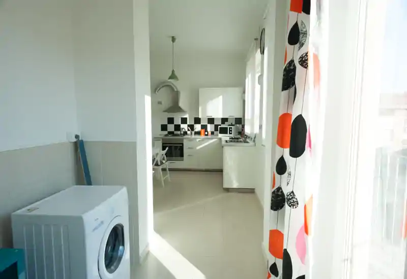 Cucina moderna e luminosa con mobili bianchi, paraschizzi a scacchi nero e bianco, forno incassato e un piccolo angolo pranzo. Ampia finestra/porta lascia entrare molta luce naturale; la tenda colorata aggiunge calore visivo.