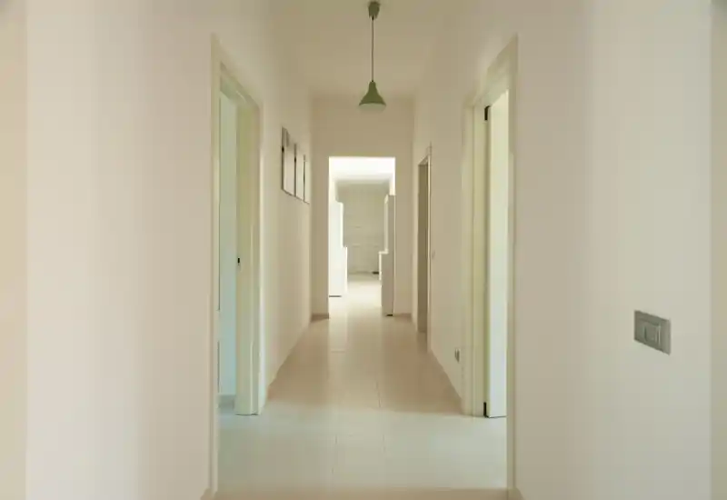 Bright, minimal hallway with white walls and tiled floor leading to other rooms; simple pendant light and neutral decor create a clean entrance view.