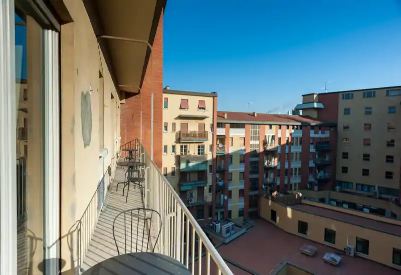 Balcone soleggiato e stretto con ringhiera in metallo e piccoli tavoli, affacciato su palazzi vicini — perfetto per un caffè mattutino con vista sulla città.