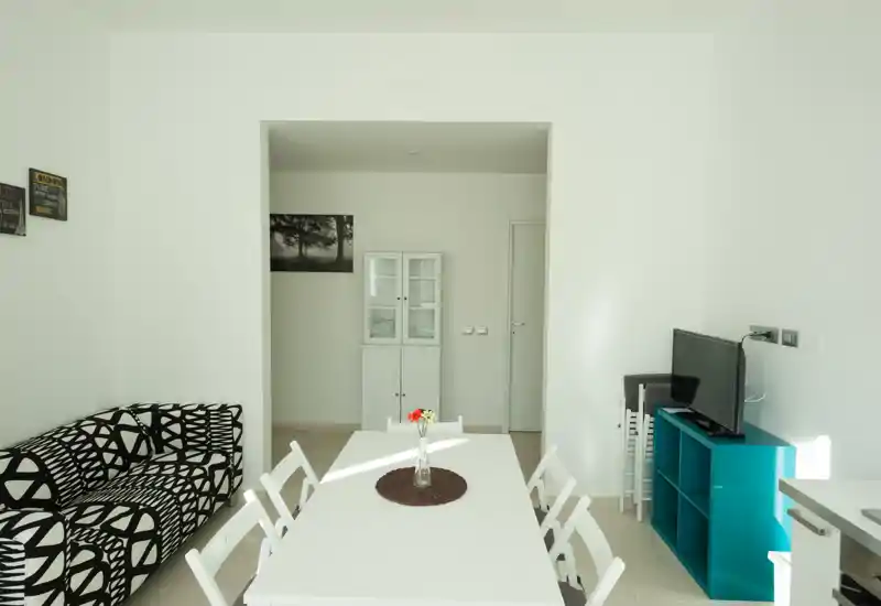 Zona pranzo luminosa e moderna con un lungo tavolo bianco, sedie pieghevoli e un centrotavola decorativo; aperta su un angolo living con divano a fantasia e mobile TV compatto.