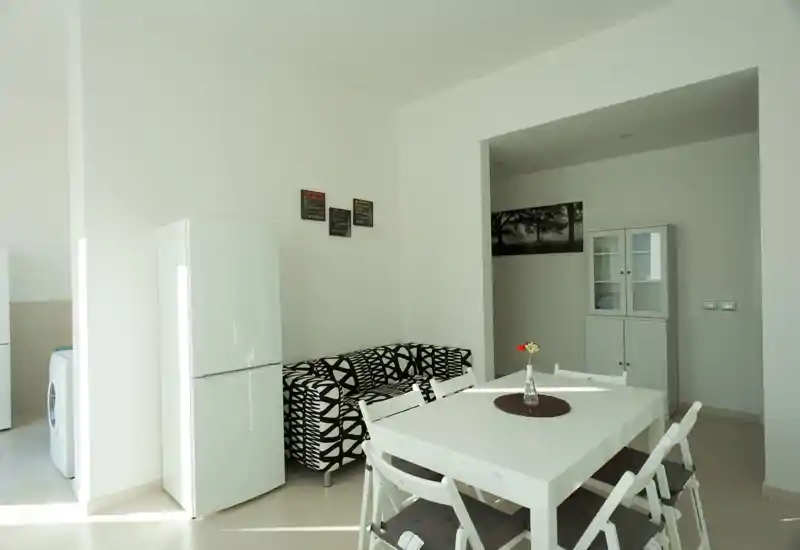 Zona giorno luminosa e minimale con tavolo da pranzo bianco, sedie pieghevoli e divano a due posti dal motivo grafico; ambiente moderno e ordinato con luce naturale.