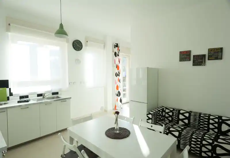 Zona giorno luminosa e moderna con angolo cottura aperto, tavolo da pranzo bianco e divano decorato — perfetta per un appartamento compatto. Finestre ampie garantiscono molta luce naturale e uno stile minimal pulito.