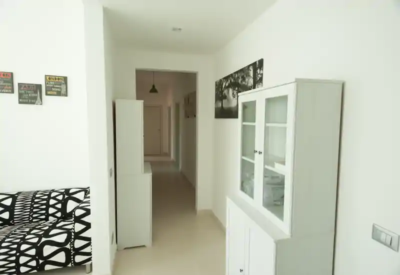 Zona giorno luminosa e minimale con divano a motivo, mobili bianchi con ante in vetro e quadri alle pareti; toni neutri e corridoio lungo visibile sullo sfondo.