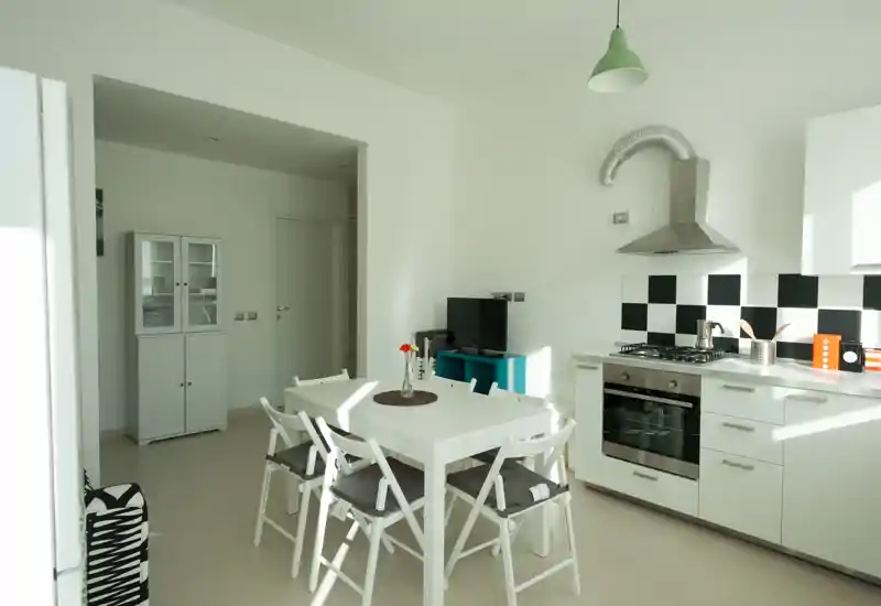 Cucina moderna e luminosa con tavolo bianco e quattro sedie, forno incassato e piano cottura a gas, paraschizzi a scacchi e dettagli decorativi essenziali. Perfetta per annunci di appartamenti compatti che valorizzano uno spazio cucina-pranzo funzionale e ben illuminato.