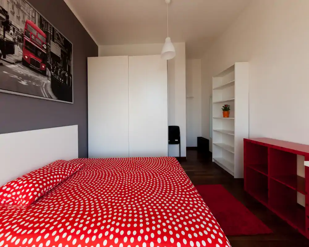 Camera da letto luminosa e di design con grande copriletto rosso a pois, armadio bianco e scaffalature aperte; pavimento in legno caldo e quadro moderno per un’atmosfera accogliente e contemporanea.
