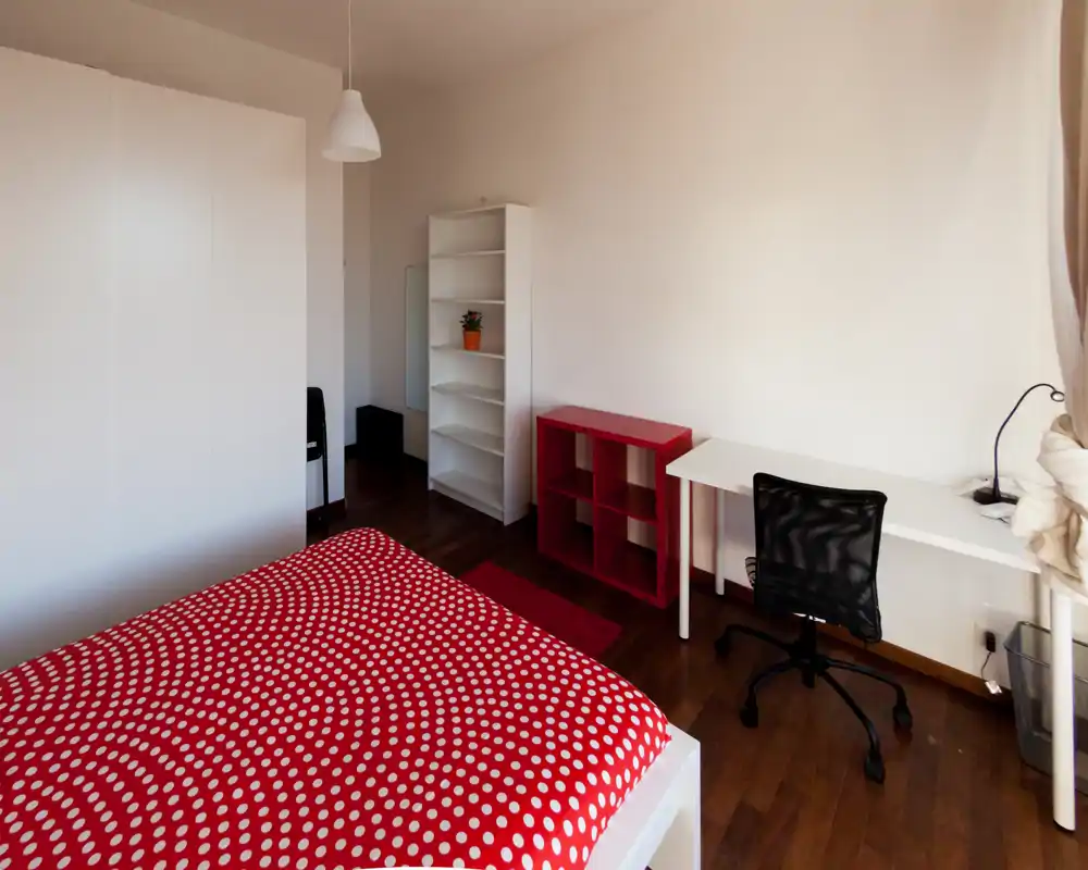 Camera da letto luminosa e ordinata con copriletto rosso a pois, armadio bianco e scrivania vicino alla finestra — ideale per uno studente o single.