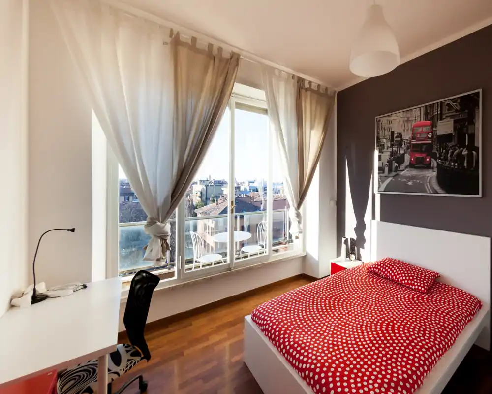 Camera da letto moderna e luminosa con letto matrimoniale e copriletto rosso a pois, ampie finestre a tutta altezza e una scrivania: ambiente arioso perfetto per soggiorni in città.
