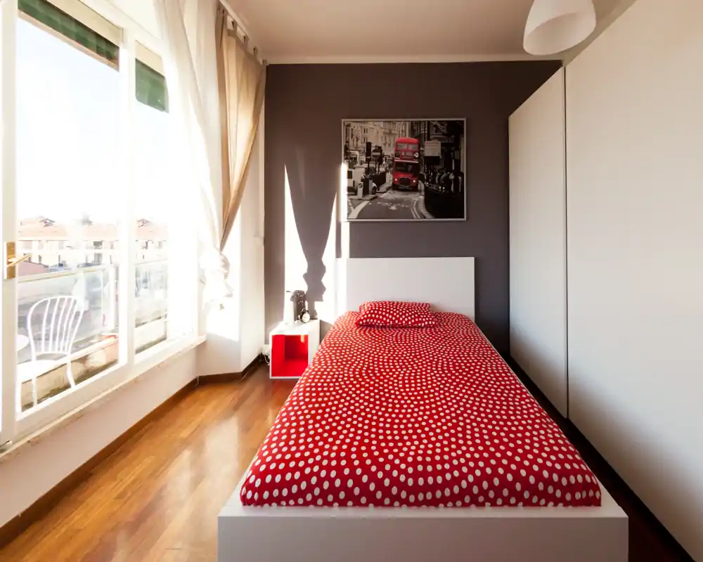 Camera singola luminosa e moderna con copriletto rosso a pois, grande finestra e accesso al balcone; linee pulite e pavimento in parquet caldo creano un ambiente accogliente.