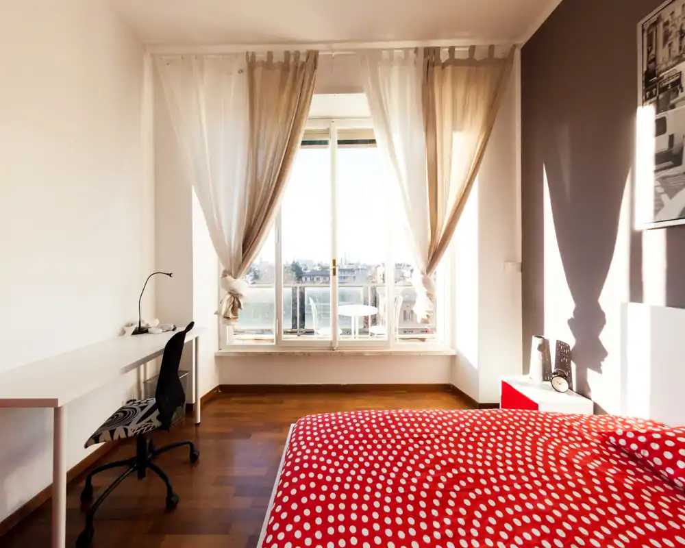 Camera da letto luminosa e moderna con copriletto rosso a pois, ampia finestra con balcone e una postazione di lavoro con scrivania e sedia.