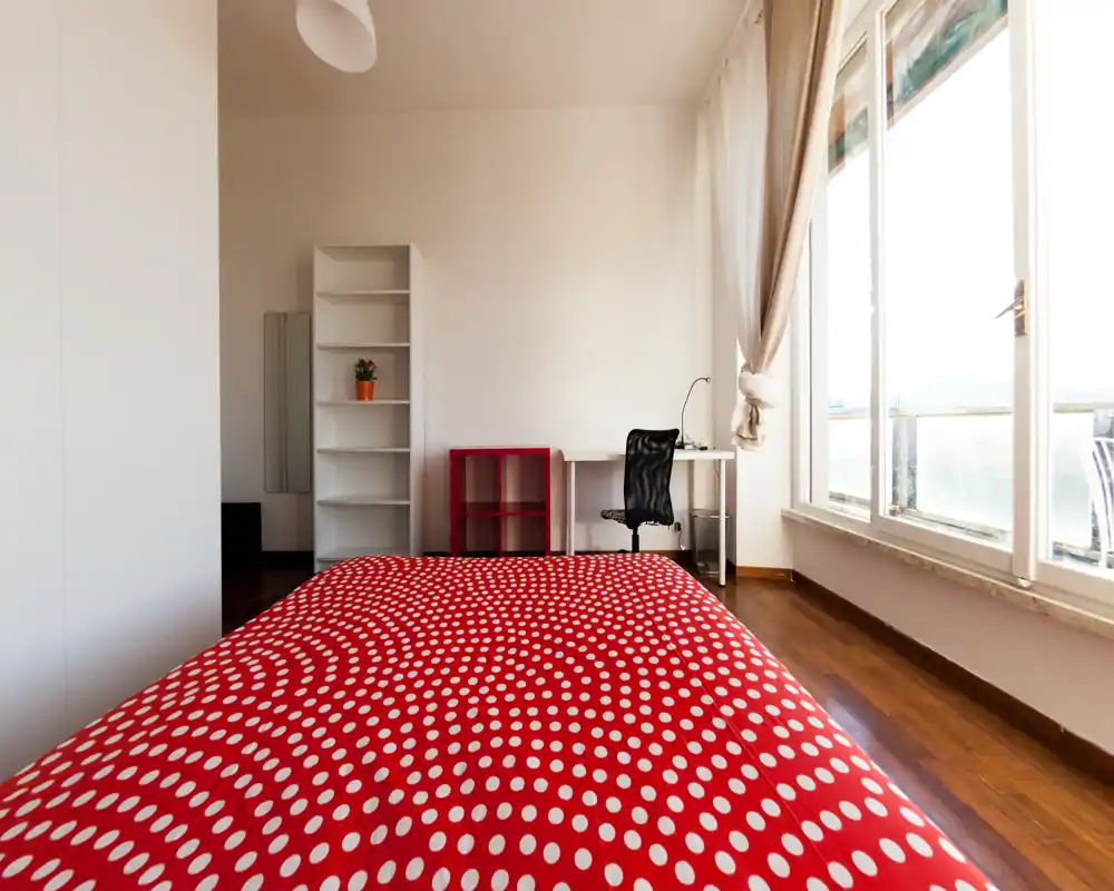 Camera da letto luminosa e moderna con copriletto rosso a pois, pavimento in legno e scrivania vicino a una grande finestra che lascia entrare molta luce naturale.