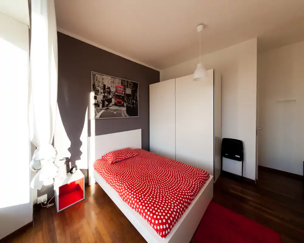 Camera da letto luminosa e curata con letto matrimoniale con copriletto rosso a pois, armadio bianco e pavimento in legno; ampia finestra per luce naturale abbondante.