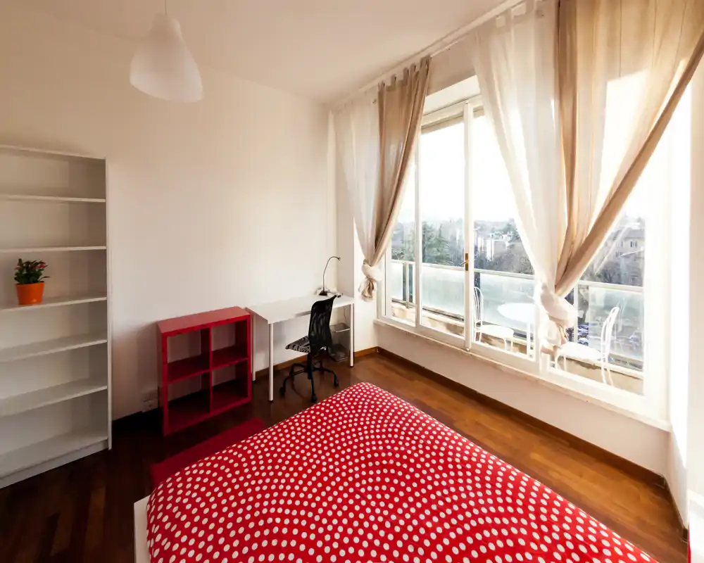 Camera da letto luminosa con grande finestra e vista sul balcone, copriletto rosso a pois, una piccola scrivania e scaffali — ambiente accogliente e ideale per rilassarsi o lavorare.