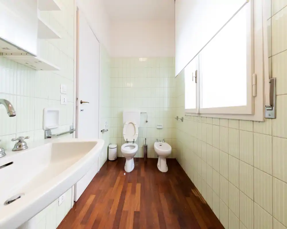 Bagno luminoso e pulito con lavabo, WC e bidet su pavimento in legno e pareti piastrellate verde chiaro. Ampia finestra per luce naturale.