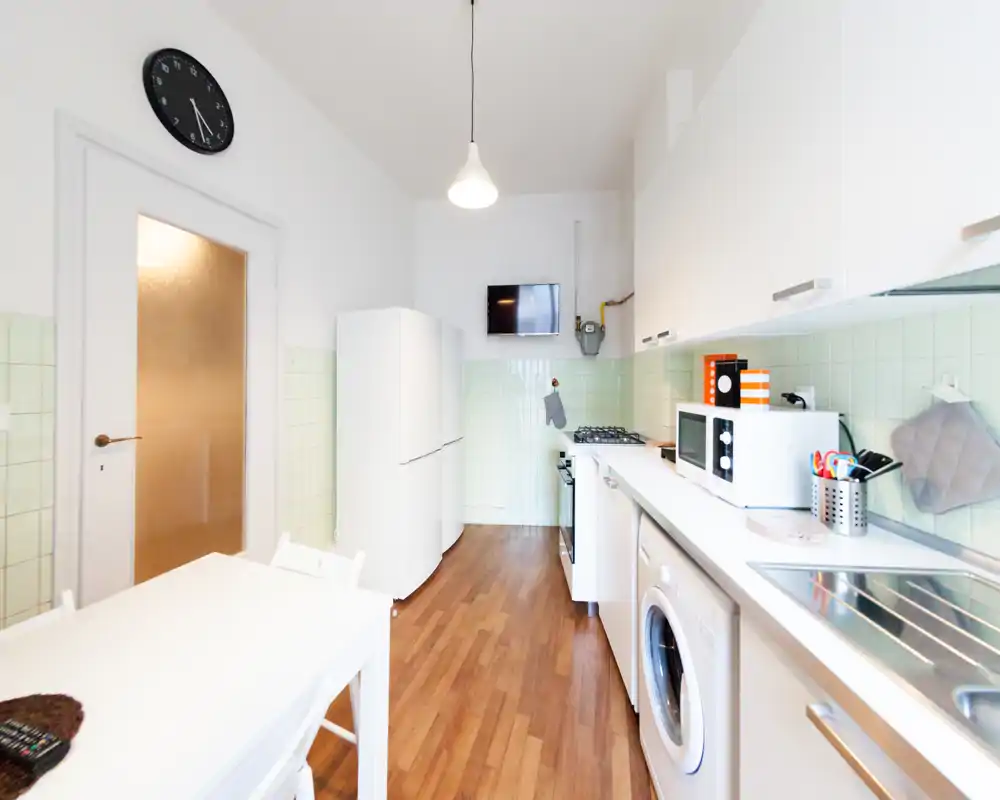 Cucina stretta e luminosa con mobili bianchi, frigorifero, piano cottura a gas, forno a microonde e lavatrice integrata; pavimento in legno caldo e piastrelle verde chiaro per un aspetto fresco e accogliente.