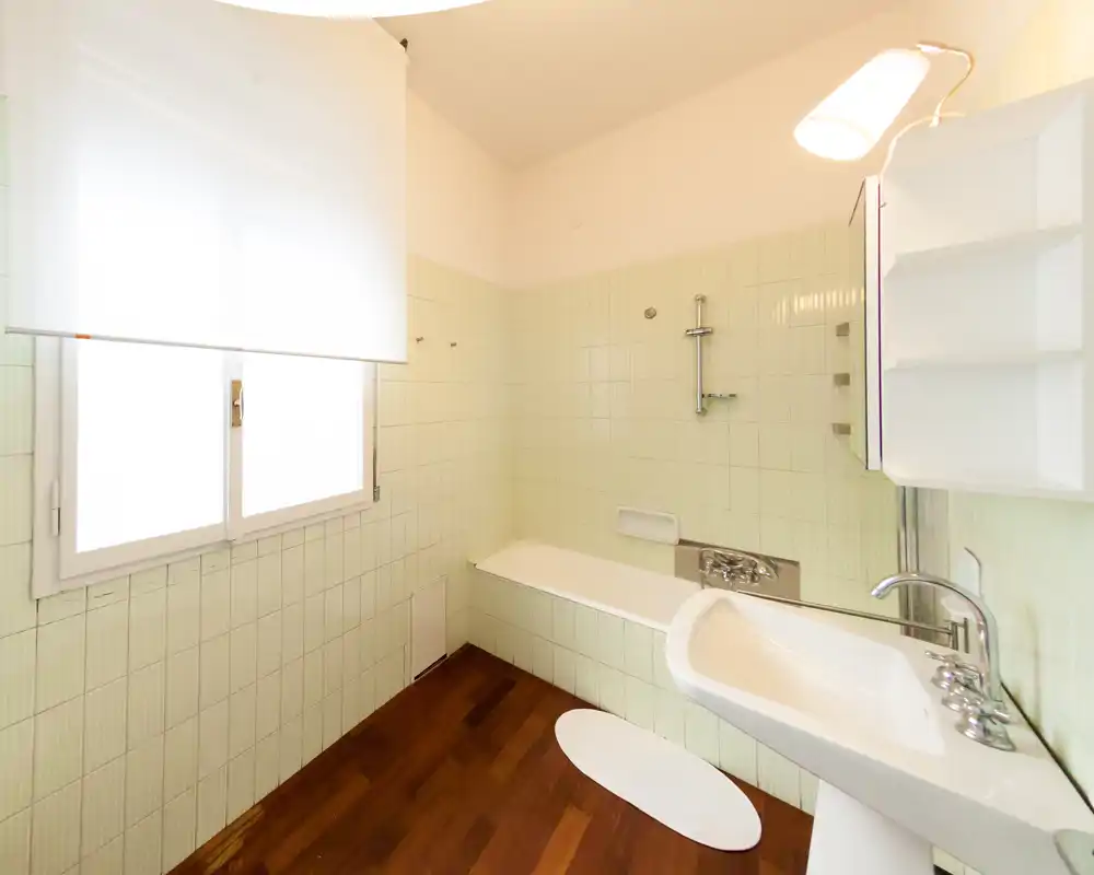 Bagno luminoso piastrellato con vasca, soffione a muro, lavabo e finestra che diffonde luce naturale. Il pavimento in legno caldo contrasta con le piastrelle verde chiaro per un aspetto pulito e minimal.