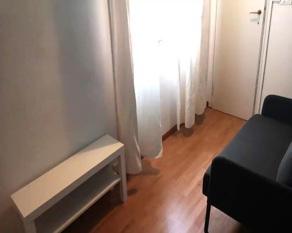 Piccola zona giorno con una poltrona imbottita scura, pavimento in legno chiaro e tende leggere che filtrano la luce. Arredamento minimale per un ambiente pratico e tranquillo.