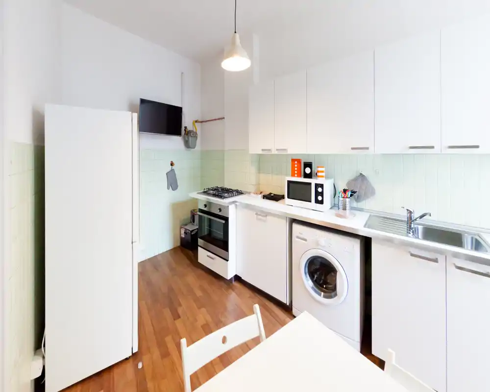 Cucina luminosa e moderna con pensili bianchi, piano cottura a gas, microonde, frigorifero e lavatrice integrata. Superfici pulite e un piccolo tavolo da pranzo per un uso quotidiano confortevole.